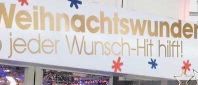 Eine Gruppe von Menschen, darunter Kinder, steht vor einem Geschäft mit einem deutschen Schild, das 'Weihnachtswunder' besagt. Sie warten wahrscheinlich in einer Schlange. Das Geschäft hat dekorative Lichter und ein Banner mit einem Stern.