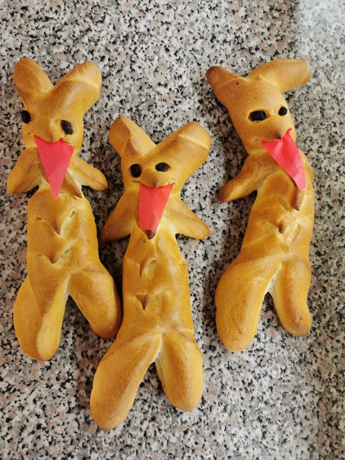 Drei goldbraune Brotfiguren in Form von Hasen liegen auf einer Marmoroberfläche. Jede Figur hat schwarze Augen und eine aus Papier gefertigte rote Zunge, die herausragt.