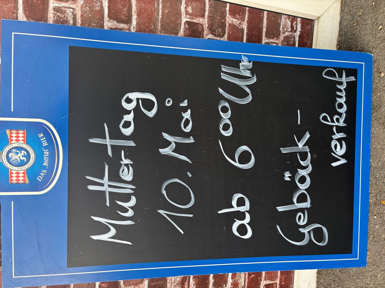 Blackboard with blue border displays 'Muttertag' and '10. Mai'. A handwritten note reads 'ab 6 Uhr' and 'Gepack' with a logo at the bottom left.