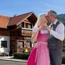 Ein Paar in traditioneller bayerischer Kleidung küsst sich vor einem Chalet mit einem grünen Schild. Bäume und Büsche sind im Hintergrund zu sehen.