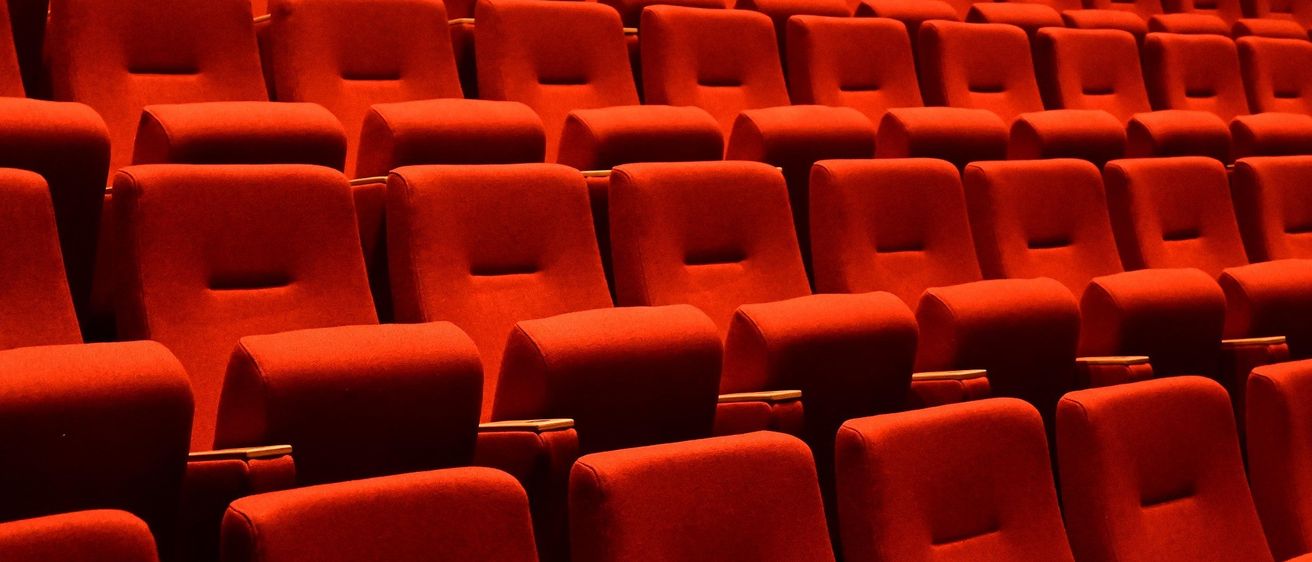 Ein Theater mit Reihen roter Samtsessel und dunklem Hintergrund. Jeder Sitz hat eine rechteckige Armlehne. Die Sitze sind ordentlich in Reihen angeordnet.