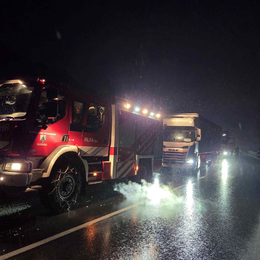 Feuerwehrfahrzeuge mit eingeschalteten Lichtern fahren bei Regen auf einer nassen Straße. Ein LKW hat Rauch, der aus dem Heck kommt.