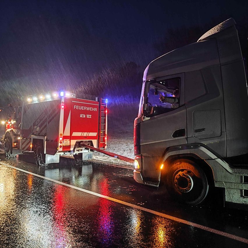 Ein Feuerwehrwagen mit blinkenden Lichtern zieht einen großen Lkw auf einer nassen Straße. Es regnet stark.