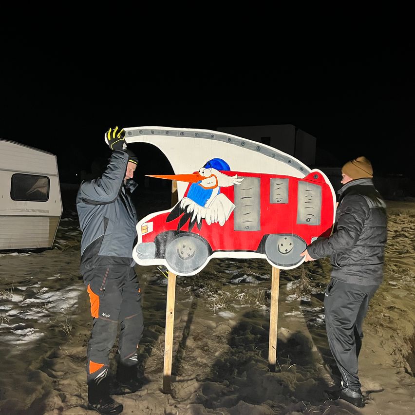 Zwei Männer halten eine große Pappaufsteller eines Wohnwagens mit einem Storch darauf. Sie sind draußen bei Nacht.