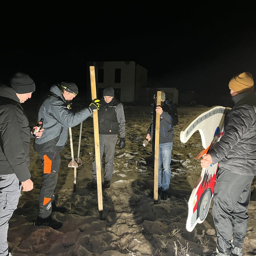 Eine Gruppe von Männern steht bei Nacht draußen und hält Holzstangen. Einer hält eine Schaufel, und ein anderer hält ein großes Schild. Sie befinden sich in einem verschneiten Bereich.