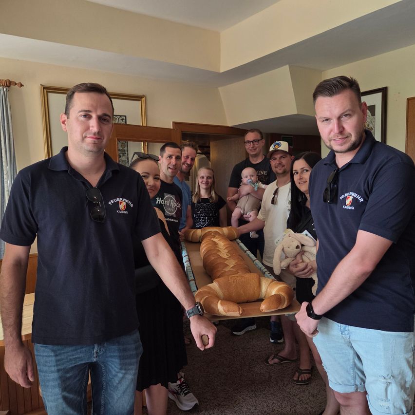 Eine Gruppe von Menschen, darunter zwei Männer in blauen Poloshirts, stehen um ein großes Brot auf einem Tisch.