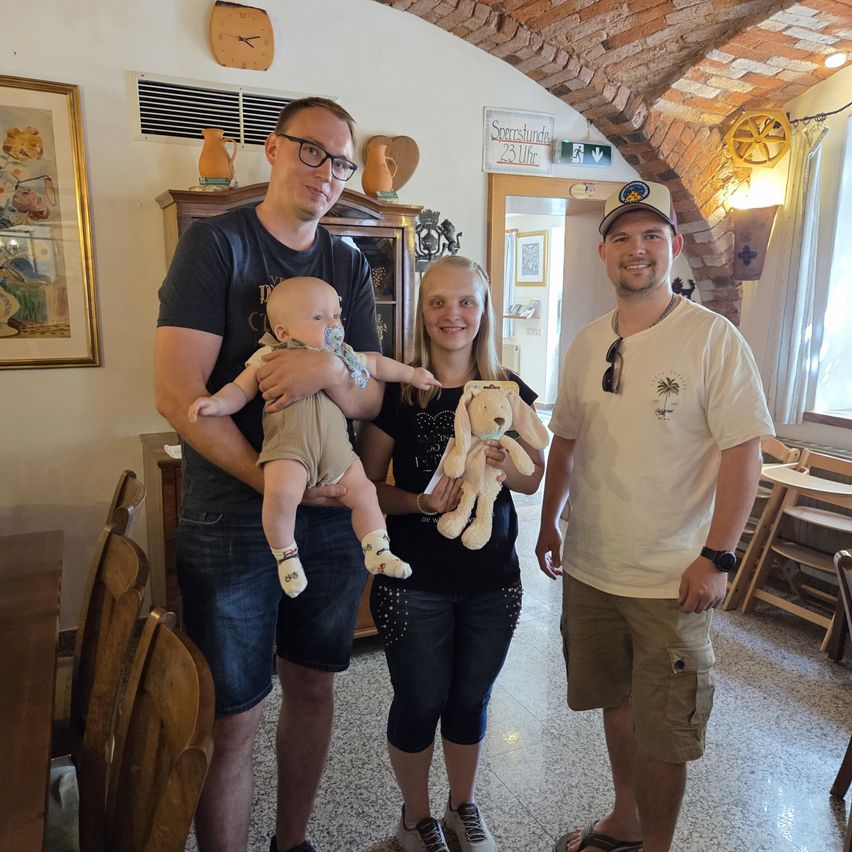 Drei Personen stehen in einem Raum mit einem Baby, einer Frau, die ein Kuscheltier hält, und einem Mann mit einer Kappe. Hinter ihnen steht ein Schrank mit dekorativen Gegenständen.