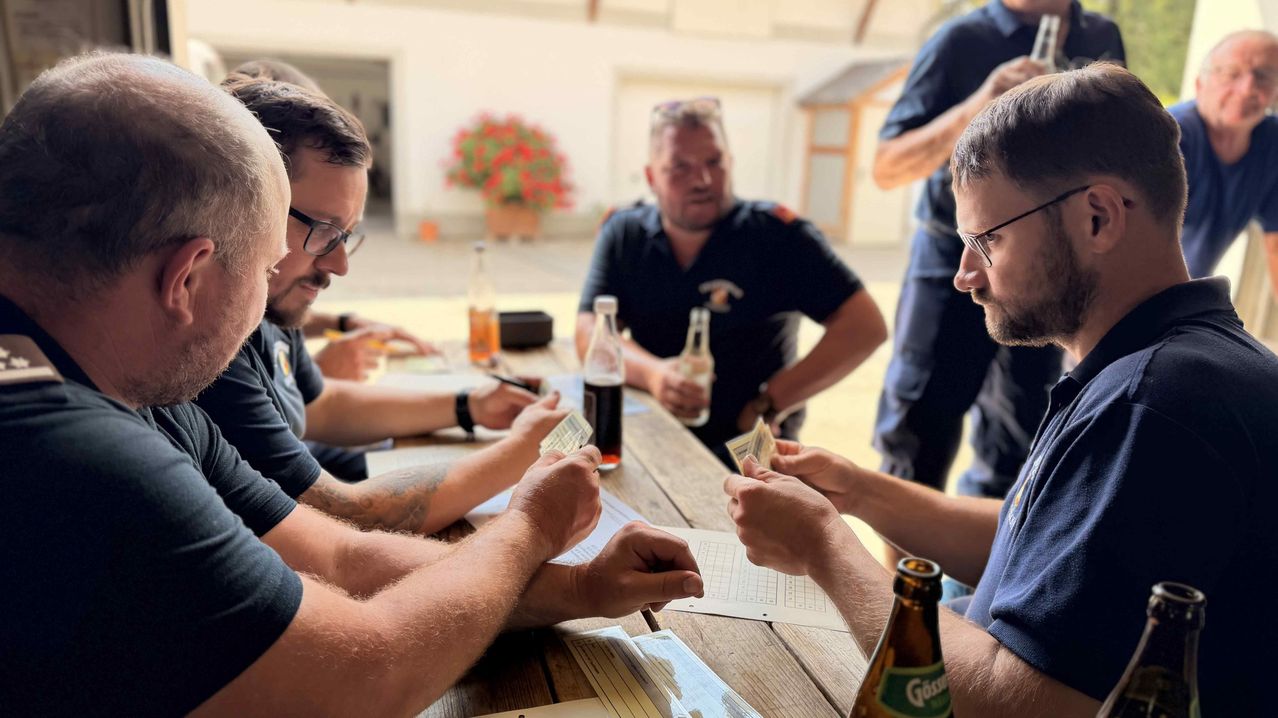 Five men sit around a wooden table, engaged in a card game. One man holds a pen and paper, while another holds a bottle. A building and a flower pot are in the background.
