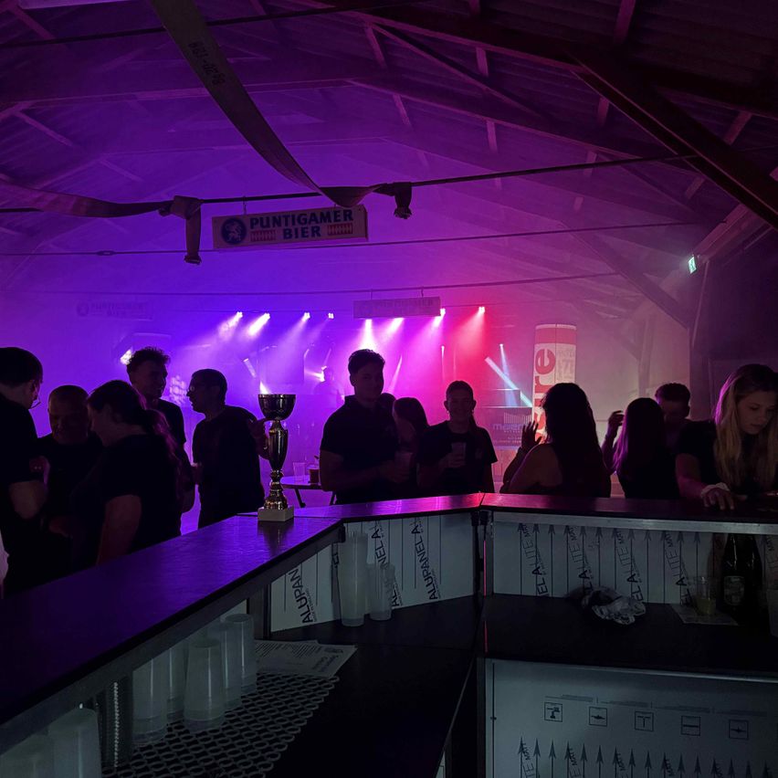 A dimly lit bar with purple lighting and a trophy on the counter, with people standing around and a sign above reading 'PUNTIGAMER BIER'.