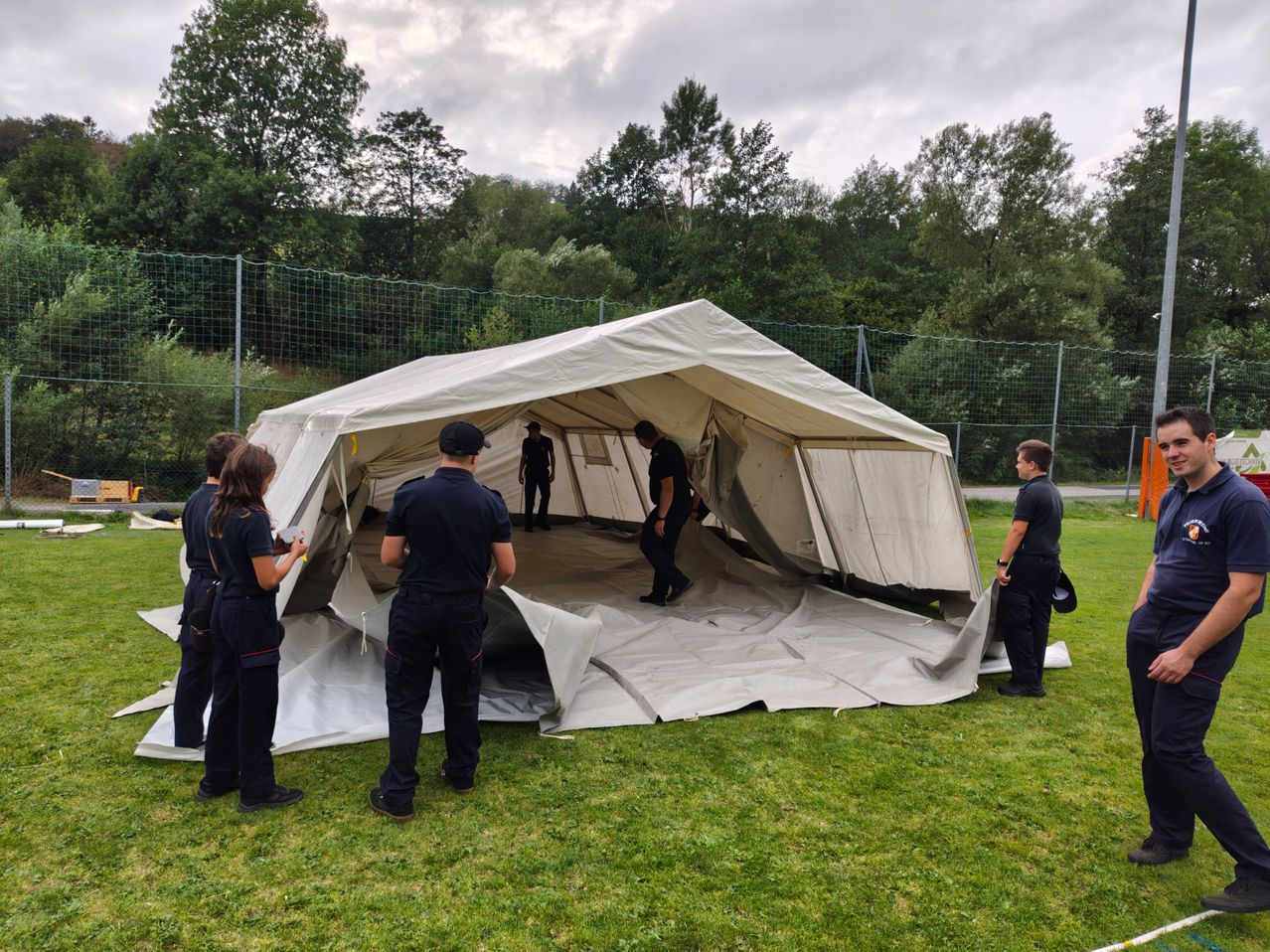 Eine Gruppe von Menschen in Uniform richtet ein großes weißes Zelt auf einem Rasenfeld ein, mit einem Zaun und Bäumen im Hintergrund.