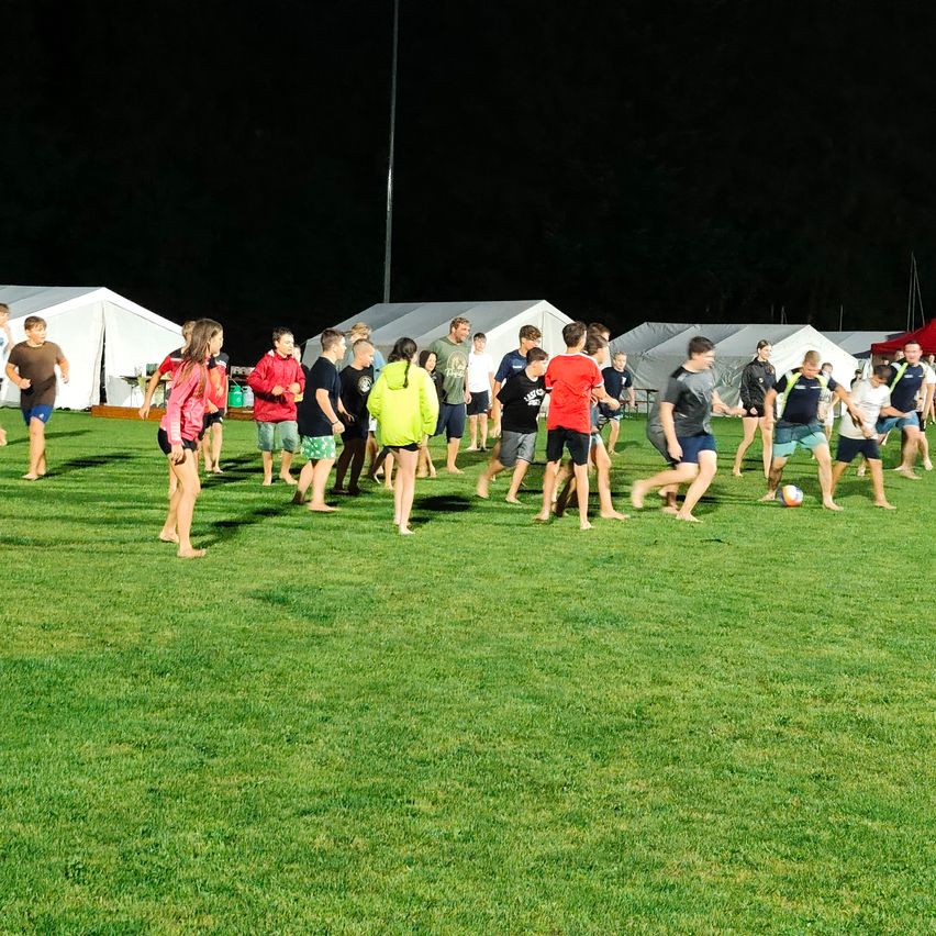 Eine Gruppe von Menschen spielt Fußball auf einem Rasenfeld bei Nacht. Einige tragen Shorts, andere lange Ärmel. Zelte und ein roter Baldachin sind im Hintergrund zu sehen.