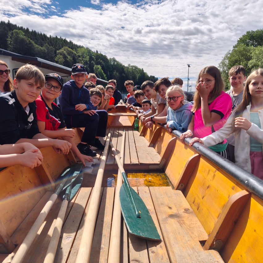 Eine Gruppe von Kindern und Jugendlichen fährt in einem hölzernen Boot auf einem Fluss, mit einem grünen Paddel in der Mitte.