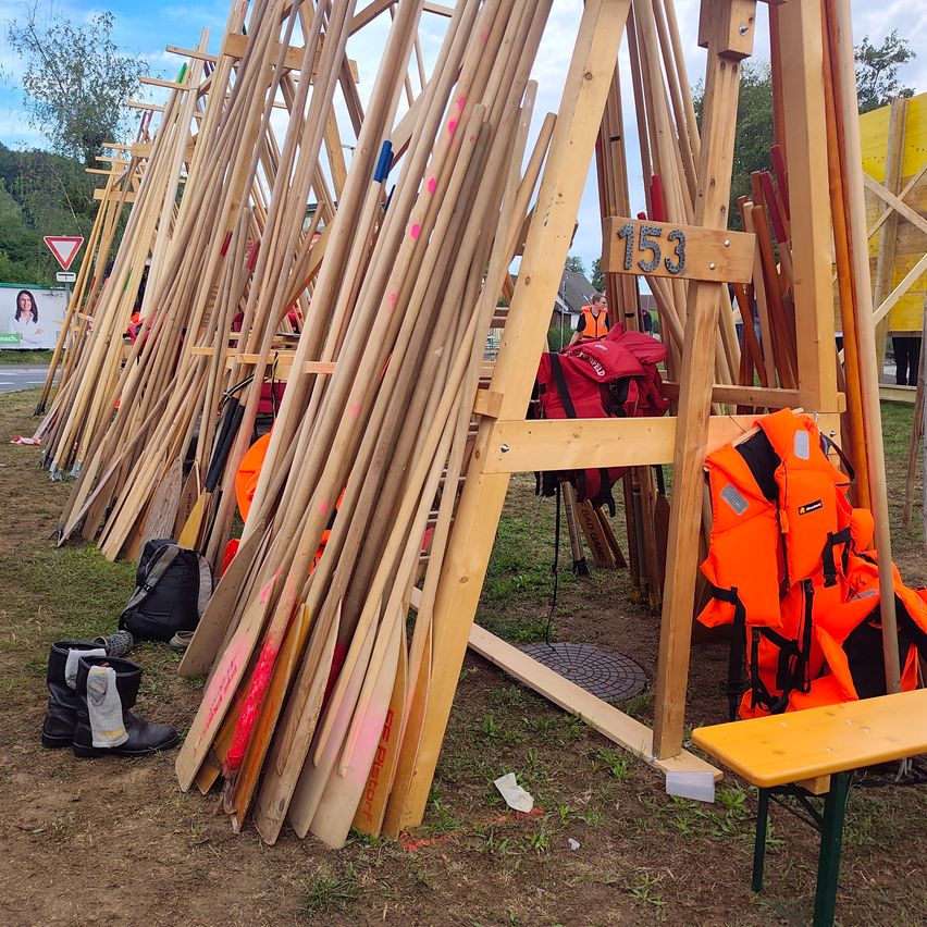Eine Reihe von Holzstangen mit Nummern und Rettungswesten, einige mit rosa Punkten, in einer Außenumgebung mit einer Bank und einem Grasbereich angeordnet.
