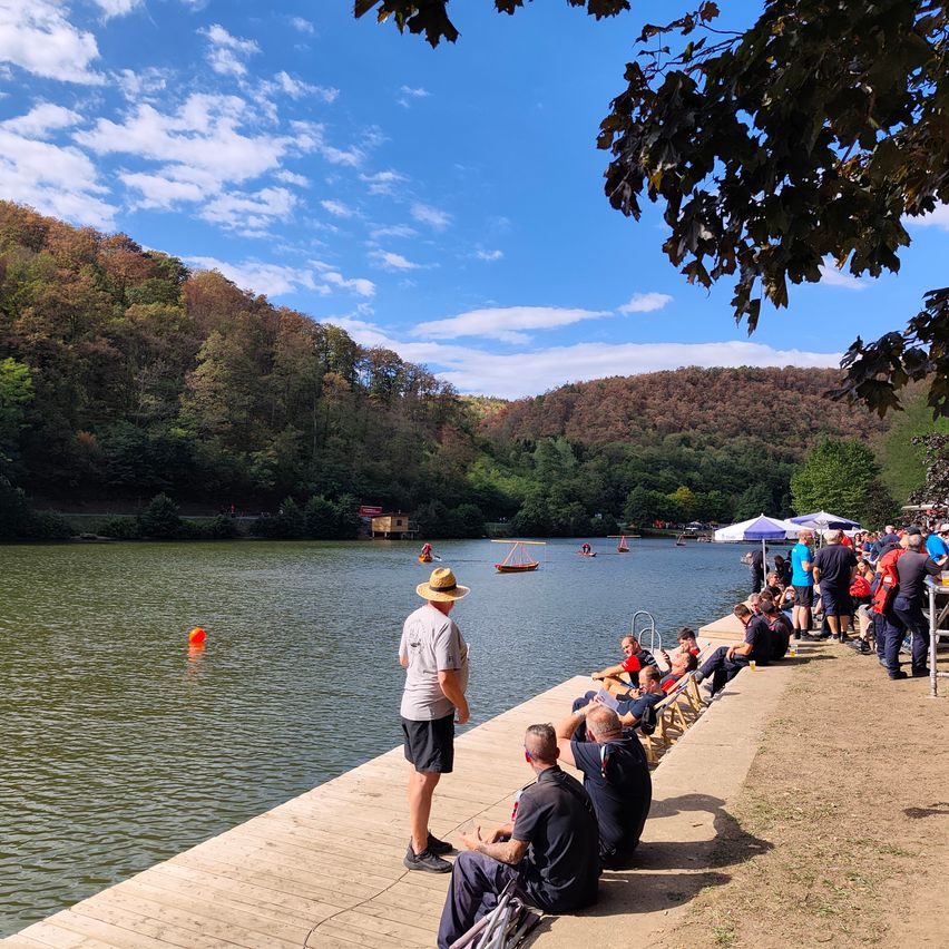 Eine Gruppe von Menschen versammelt sich auf einem hölzernen Steg am See, mit mehreren Booten auf dem Wasser. Ein Mann steht auf dem Steg, während andere auf Stühlen sitzen oder in der Nähe stehen. Der See ist von Bäumen und Bergen umgeben.