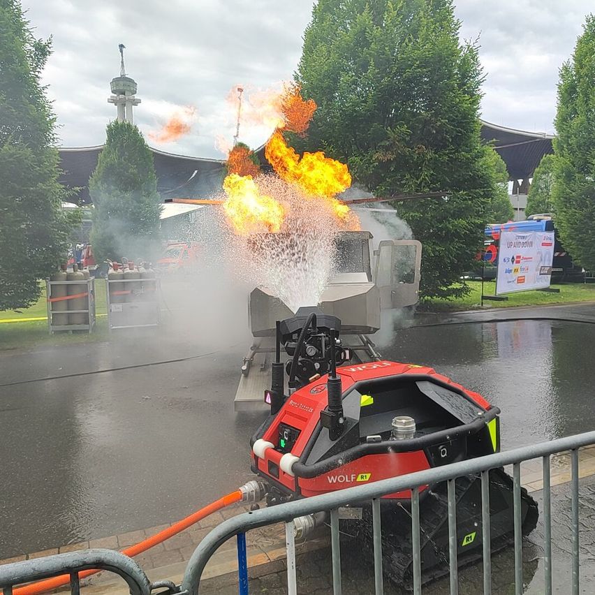 A red robot vehicle with the word Wolf on it is spraying water on a fire. There are trees and a building in the background.