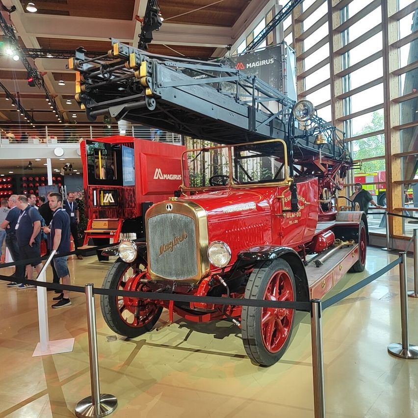 An old red fire truck with a ladder on top is on display inside a building. Many people are looking at it.