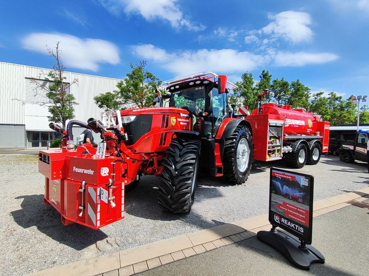 A large red tractor with firefighting equipment is parked on a road. A building and trees are in the background.