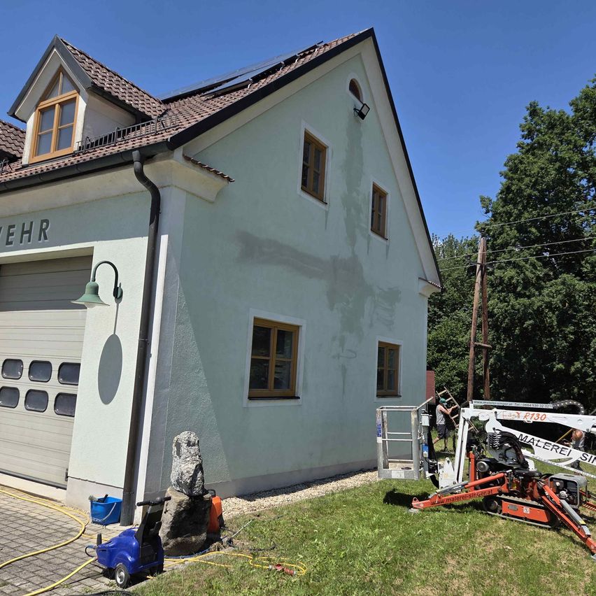 Ein Haus mit Solarmodulen auf dem Dach wird gestrichen. Eine Leiter lehnt an der Wand. Ein blauer Eimer steht auf dem Boden. Ein Maschinenfahrzeug ist vor dem Haus geparkt.