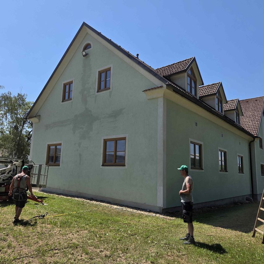 Zwei Männer arbeiten an der Außenseite eines Hauses. Einer benutzt ein Elektrowerkzeug, der andere beobachtet. Das Haus hat ein braunes Dach und hellgrüne Wände.