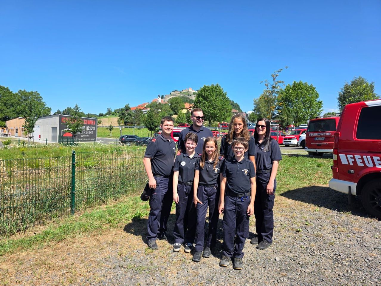 Eine Gruppe junger Menschen in Uniform steht vor einem Zaun mit einem Schild, einem Feuerwehrauto und einem roten Van im Hintergrund.