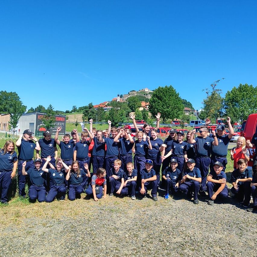Eine Gruppe junger Menschen in blauen Uniformen posiert für ein Foto mit erhobenen Händen. Sie stehen auf einer Kiesfläche mit Bäumen und einem Gebäude im Hintergrund.