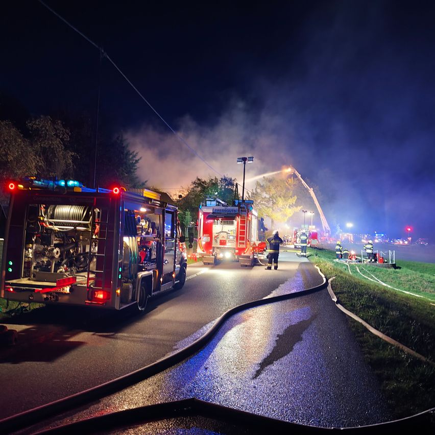 Feuerwehrleute reagieren nachts auf einen Brand, mit Feuerwehrwagen und einer Leiterwagen am Einsatzort. Rauch steigt aus einem Gebäude auf, und Notfalllichter blinken.