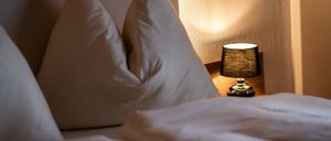 Ein ordentlich gemachtes Bett mit zwei weißen Kissen und einer Lampe auf dem Nachttisch. Der Raum ist sanft beleuchtet.