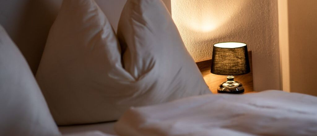 Ein ordentlich gemachtes Bett mit zwei weißen Kissen und einer Lampe auf dem Nachttisch. Der Raum ist sanft beleuchtet.