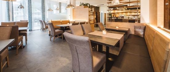 A modern restaurant with tiled floors, large tables, and gray chairs. A wooden counter holds shelves with plants and cups, and pendant lights hang from the ceiling.