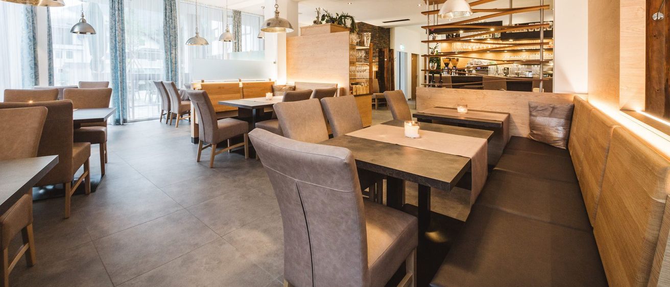 Ein modernes Restaurant mit gefliesten Böden, großen Tischen und grauen Stühlen. Ein Holztresen mit Regalen, Pflanzen und Tassen, und Pendelleuchten hängen von der Decke.