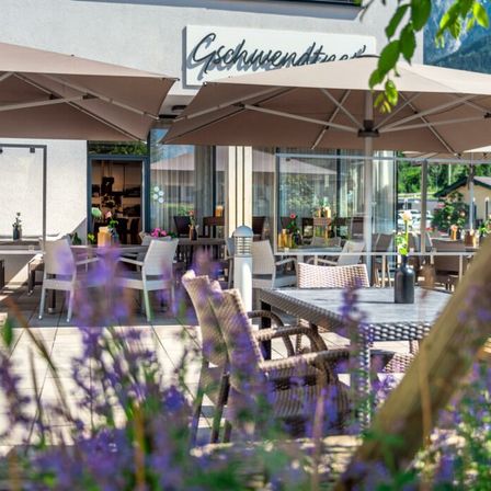 Außenbestuhlung in einem eleganten Restaurant mit großen Sonnenschirmen, umgeben von lila Blumen und Bergen im Hintergrund.