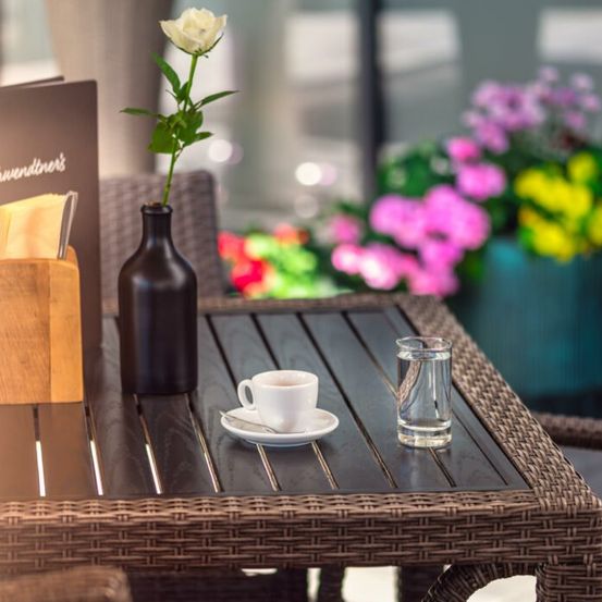Ein brauner Korbtisch mit einer weißen Tasse, Untertasse und einer Vase mit einer weißen Blume sowie einem Glas Wasser.