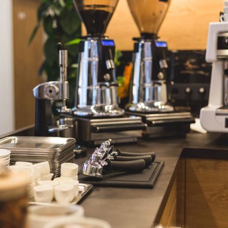 Eine Kaffeestation mit mehreren Kaffeemaschinen, einem Stapel Teller und Tassen. Drei schwarze Kaffeemühlen mit Digitalanzeigen und silbernen Griffen.