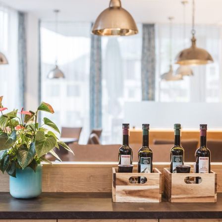 Ein Holztresen mit einer blauen Vase mit einer Pflanze und vier Weinflaschen in Holzkisten darauf. Dahinter ist ein Essbereich mit mehreren Stühlen und Lampen zu sehen.
