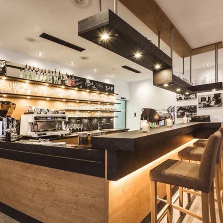 Ein modernes Café-Interieur mit einer Holztheke, Stühlen und einer Espressomaschine. Regale halten verschiedene Flaschen und Gläser. Die Decke ist mit Holzbalken und Lichtern ausgestattet.