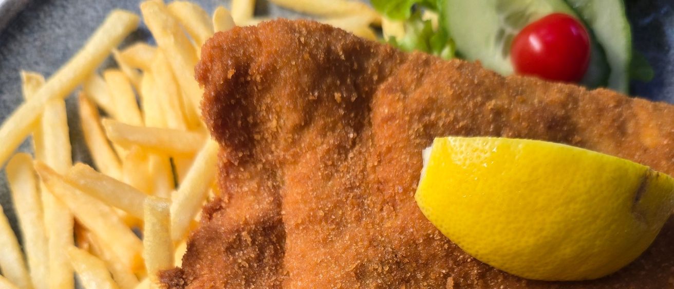 Ein Teller mit gebratenem Schnitzel, einer Zitronenscheibe, Pommes und einer Gurkenscheibe.