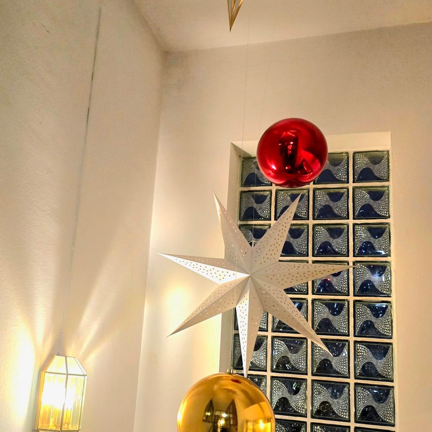 Ein weißer Stern hängt von der Decke in der Nähe eines blauen Mosaikfensters. Darunter hängen zwei glänzende Kugeln, eine rot und eine gold.