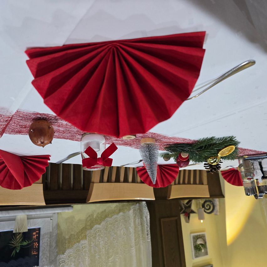 Eine Tischdekoration mit roten Servietten, einem roten Band, einem Weihnachtsbaum und Ornamenten. Die Szene reflektiert sich an einer Decke mit einer gelblichen Wand und Vorhängen.