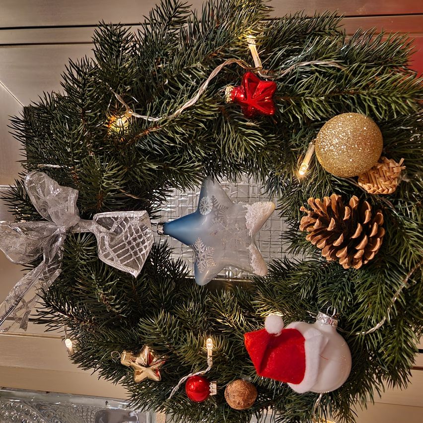Ein Kranz mit einem roten Stern, einem blauen Stern, einer goldenen Kugel und einem Weihnachtsmannhut. Er hat ein silbernes Band und Tannenzapfen. Der Kranz steht auf einer Holzoberfläche.