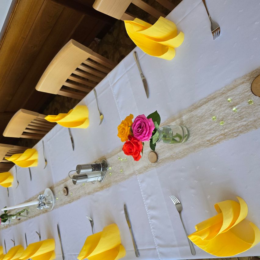Ein langer Esstisch mit weißer Tischdecke, gedeckt mit gelben Servietten, Besteck und einem Blumencenterpiece in verschiedenen Farben. Jede Tischplatzierung beinhaltet eine Gabel, ein Messer und einen Löffel.