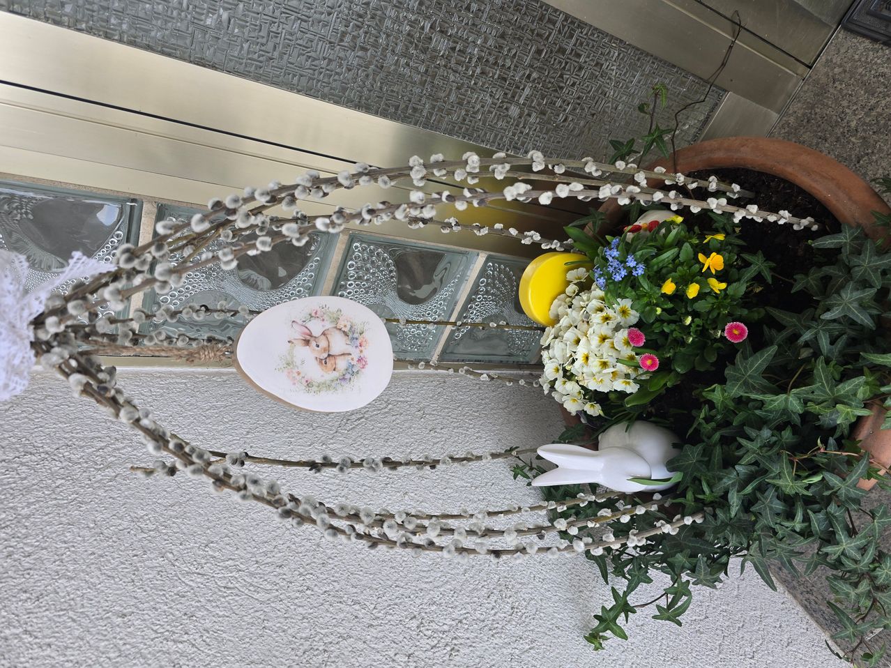 Eine dekorative Osterdekoration mit einer weißen Hasenfigur, einem Osterei und Frühlingsblumen in einem Topf. Die Dekoration steht vor einem Hintergrund aus weißen und grauen Fliesen.