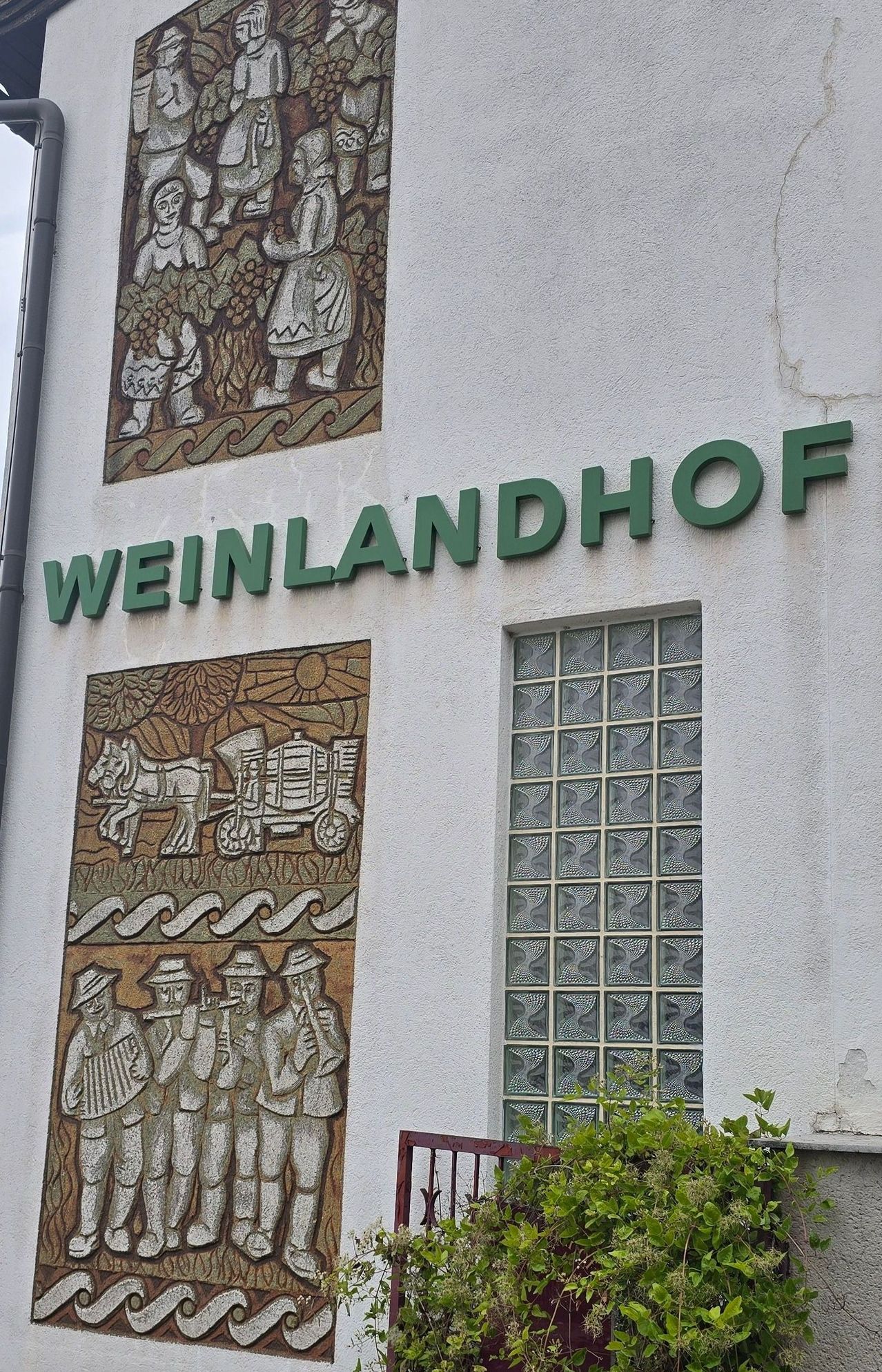 Ein weißes Gebäude mit einem Schild, das 'Weinlandhof' besagt. Zwei Wandgemälde zeigen Szenen von Menschen, die Trauben ernten, und einen von Pferden gezogenen Wagen. Ein Fenster mit rautenförmigen Glasscheiben ist sichtbar.