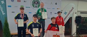 Vier Jungen stehen auf einer Bühne und halten Medaillen und Urkunden. Ein Junge hält eine Flagge mit einem Wappen.