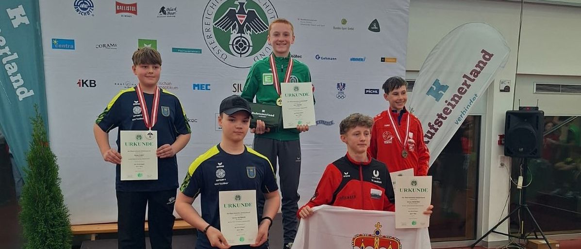 Vier Jungen stehen auf einer Bühne und halten Medaillen und Urkunden. Ein Junge hält eine Flagge mit einem Wappen.