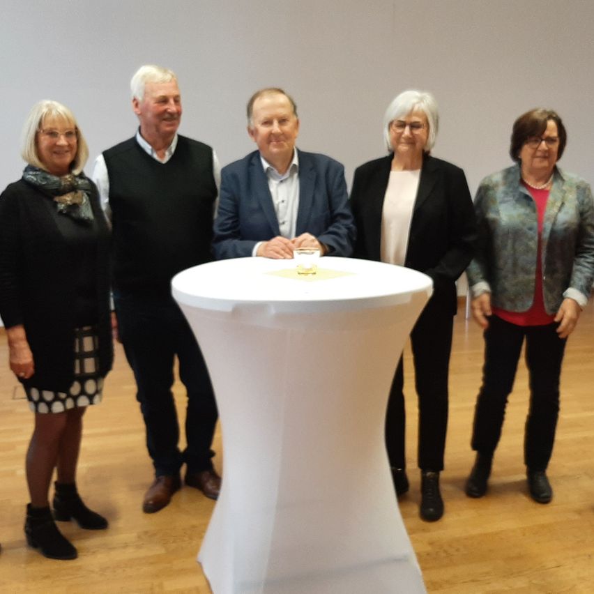 Fünf Personen stehen neben einem runden Tisch. Sie scheinen für ein Foto zu posieren. Sie alle lächeln.