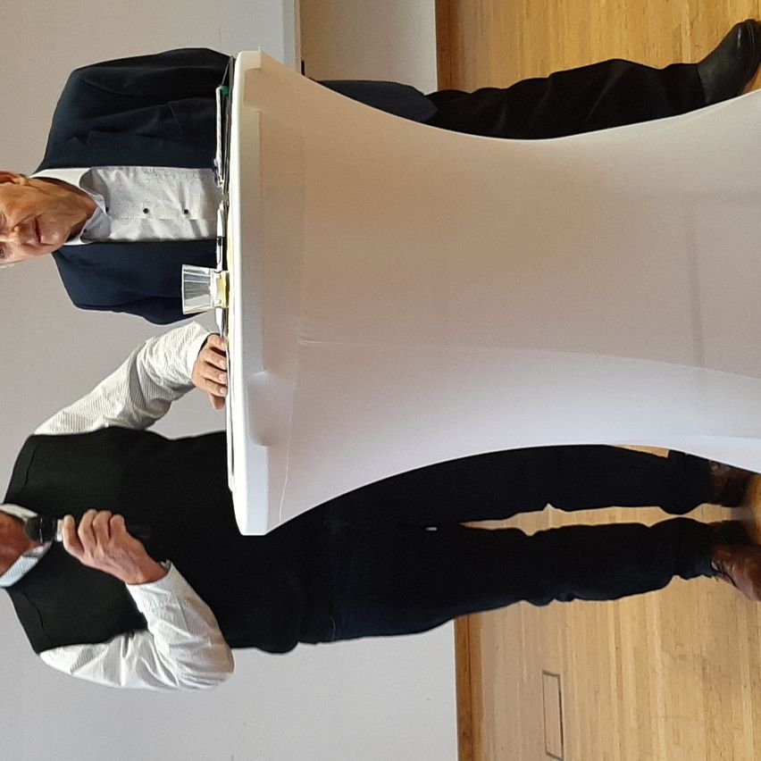 Zwei Männer, einer steht an einem Rednerpult, der andere lehnt sich darüber, beide sind in formeller Kleidung gekleidet. Der Ort scheint eine Konferenz oder Präsentation zu sein.