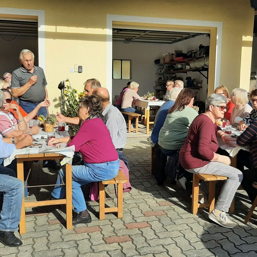 Eine Gruppe älterer Menschen versammelt sich um Tische vor einem Gebäude, einige sitzen auf Bänken, andere stehen. Ein Mann steht und gestikuliert, während andere in Gespräche vertieft sind.
