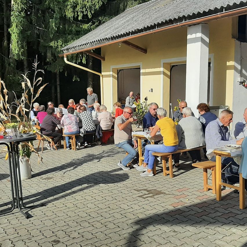 Eine Gruppe älterer Menschen sitzt um Tische in einem Außenbereich. Ein Gebäude mit gelben Wänden und einem grauen Dach ist im Hintergrund zu sehen.