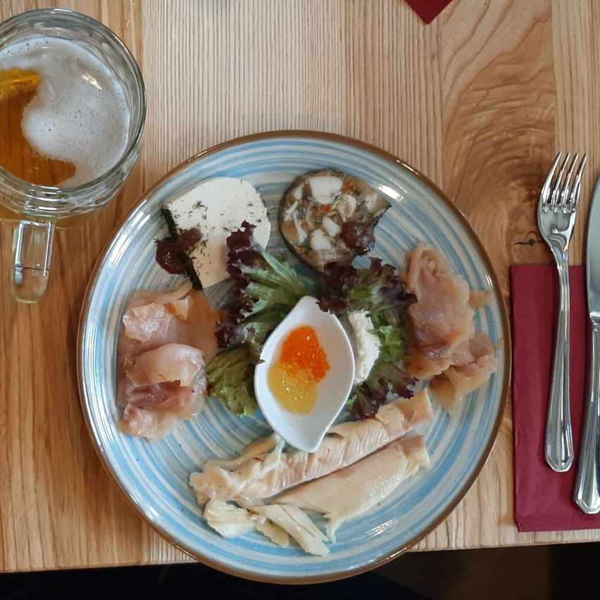 Ein blau-weißer Teller mit einer Mischung aus Fisch, Käse und Salat steht auf einem Holztisch. Ein Glas Bier steht links, und Silberbesteck ist rechts.