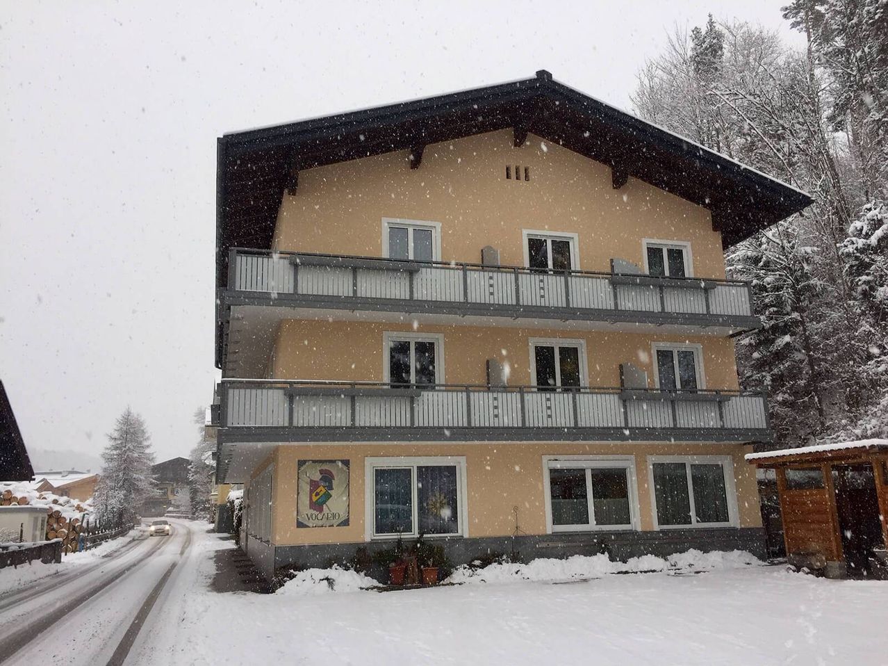 Ein mehrstöckiges Gebäude mit Balkonen und Topfpflanzen davor, im Schnee bedeckt, an einer verschneiten Straße mit einem vorbeifahrenden Auto.